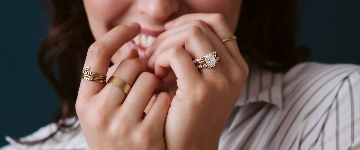 Woman smiling and covering her face with multiple gold stacking rings, a diamond engagement ring and wedding band.